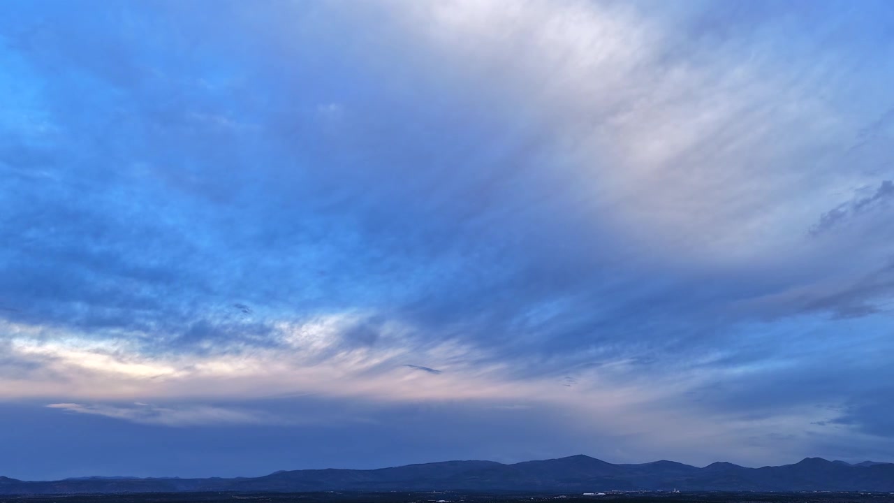 Timelapse udara lanskap pegunungan yang tenang. Awan yang menyenangkan dan menyebar bergerak perlahan di atas saat cahaya senja sekitar bergeser, memancarkan warna lembut yang berubah di seluruh medan.