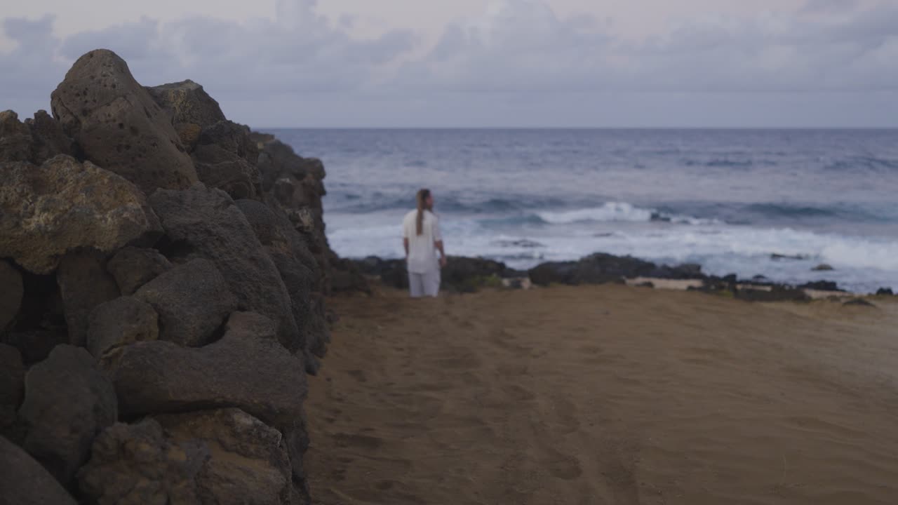 A man dressed in white walks away from the camera on a sandy coastal path beside a rugged stone wall, heading toward the ocean waves and distant horizon