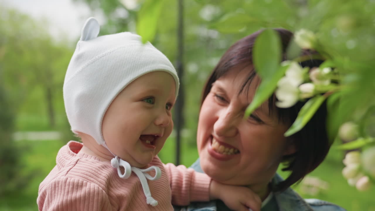 Grandmother And Infant, Senior Female Figure Playing With Infant Amidst Blooming Branches In Nature, Caucasian Grandmother And Her Baby Engaging With Lush Flowering Tree In Joyful Outdoor Scene