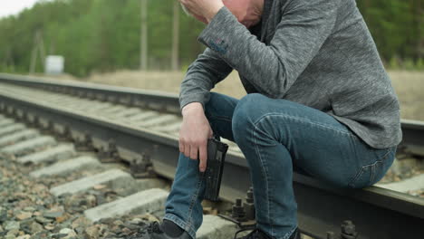 a close view of a man in a gray jacket, canvas and blue jeans, holding a pistol, sitting on a railway track, his head is bowed, and one hand is on his head, with a blurred view of a green forest