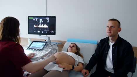 young caucasian couple is having ultrasonic examination. pregnant woman lies on the couch and her husband sits beside.