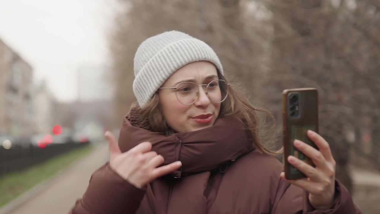 Nuori nainen kävelee. Hän keskustelee livenä kadulla. Hymyilevä nainen puhelimen kanssa kaupungin jalkakäytävällä. Valkoinen nuori nainen käy live-keskustelua kävellessään rakennusten ohi kylmällä säällä.