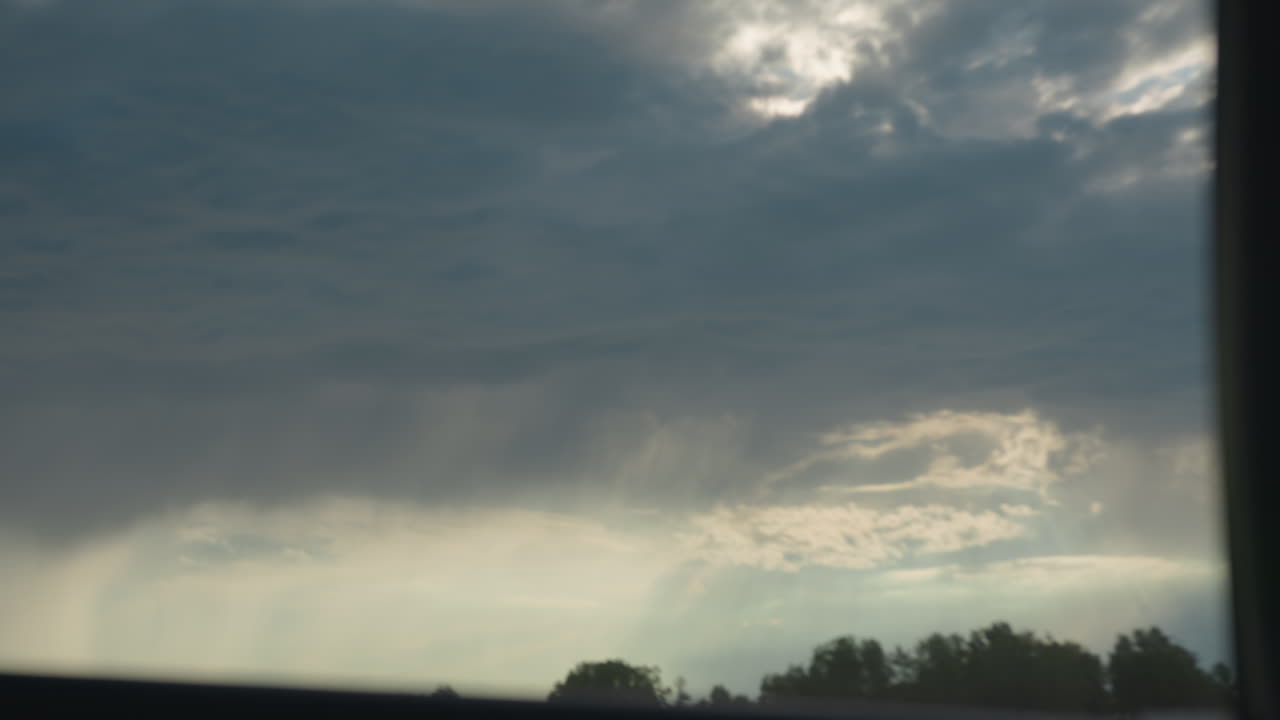 dramatic gray clouds swirling overhead, sunbeams piercing cloud base, distant tree line silhouette under expansive sky, hint of rain shafts pouring onto green canopy, serene nature scene before storm