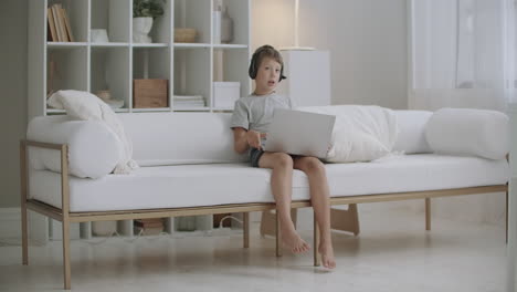 little-boy-is-chatting-with-parents-online-by-laptop-with-headphones-from-summer-camp-sitting-on-bed-at-room