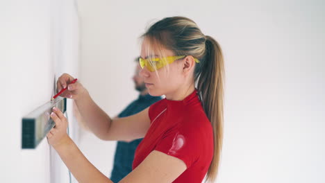 girl draws line on wall with level against man in light room