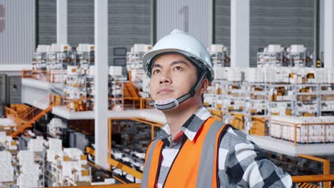 close up side view of asian male engineer with safety helmet in the warehouse with shelves full of delivery goods. standing with arms akimbo looking around, checking the stock on racks