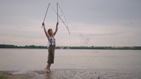 una joven hippie en un vestido y con plumas en la cabeza hace enormes burbujas de jabón al atardecer en la orilla de un lago en cámara lenta