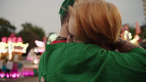 una foto en primer plano de una chica con cabello verde corto en una camisa verde a cuadros y una chica rubia con un corte de pelo corto en una camiña rosa a cuadros abrazándose y mirándose durante su cita en el parque de atracciones