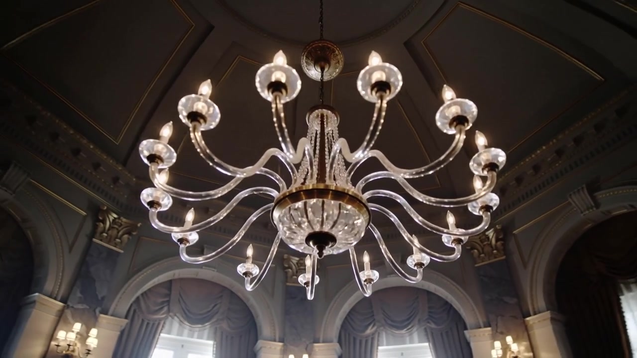 Inizio inclinazione e zoom, la telecamera inquadra un lampadario di cristallo in una sala formale, enfatizzando il soffitto.