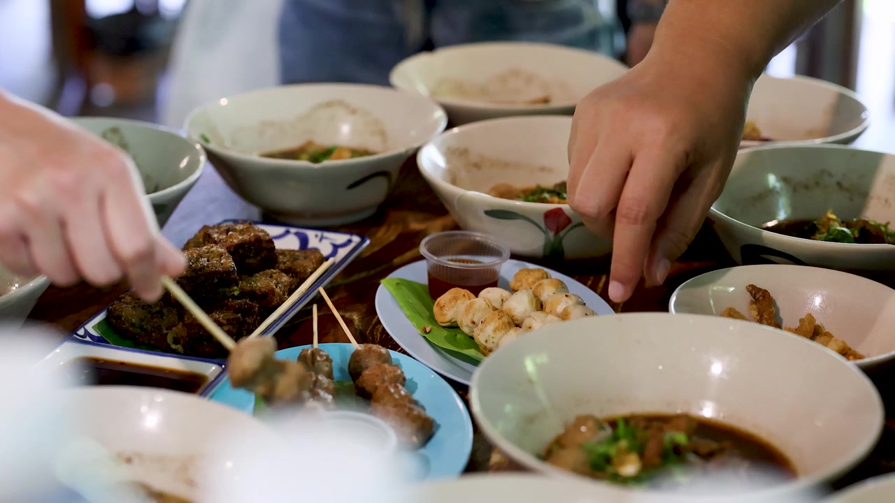 Close-up tangan mengambil sate bakso bakar di antara beberapa mangkuk sup mi Thailand