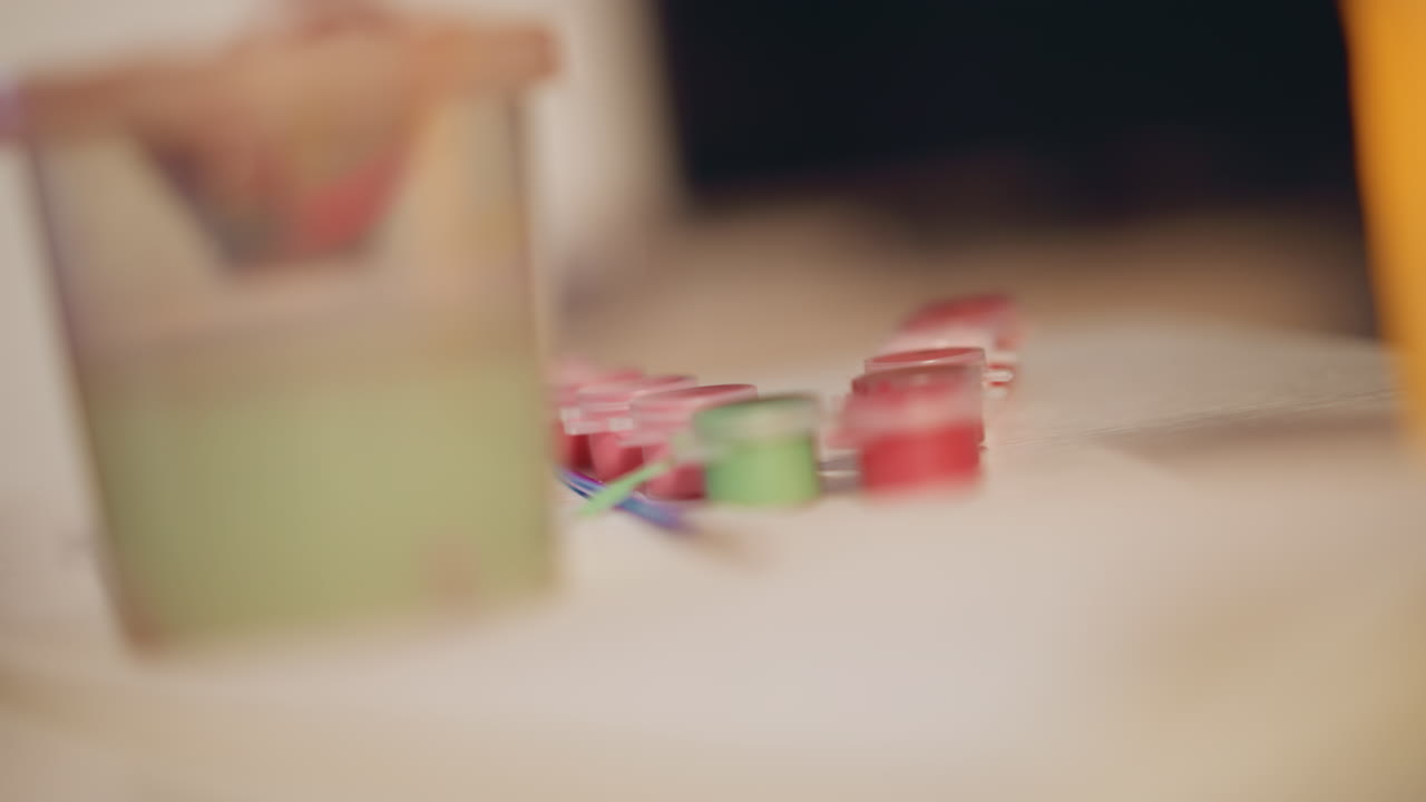 Shallow Focus Lined Paint Pots, Row Of Miniature Color Jars Beside Green Cup And Warm Candle, Soft Foreground Blur And Cinematic Shallow Depth, Tidy Workspace Before Painting Session