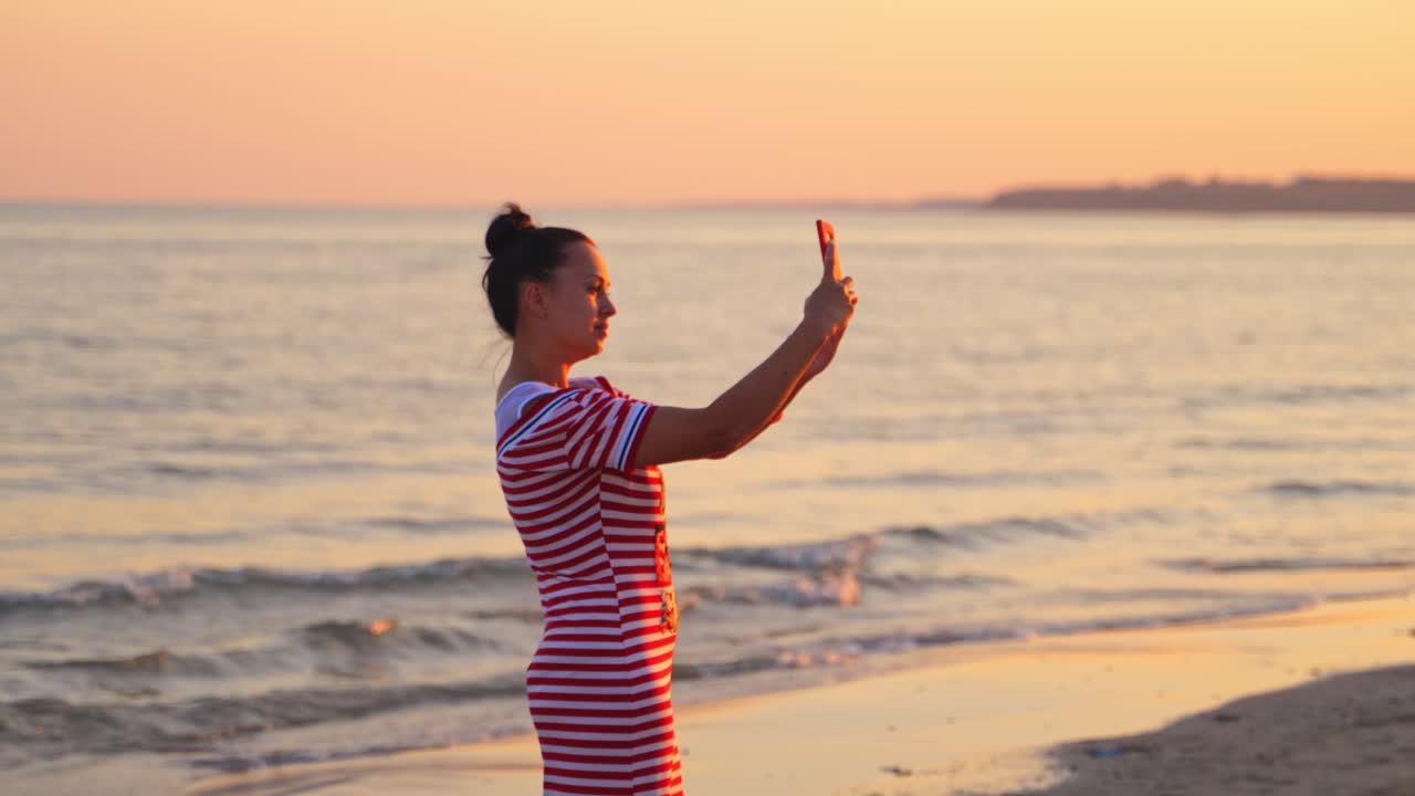 Woman takes picture of beautiful sunset sea with phone. Girl in dress holds smartphone making photo memories of summer vacation with mobile camera.