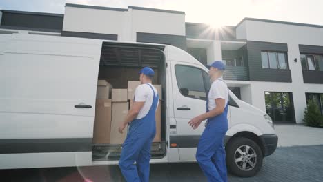 two young workers of removal company unload boxes and furniture from minibus into customer's home