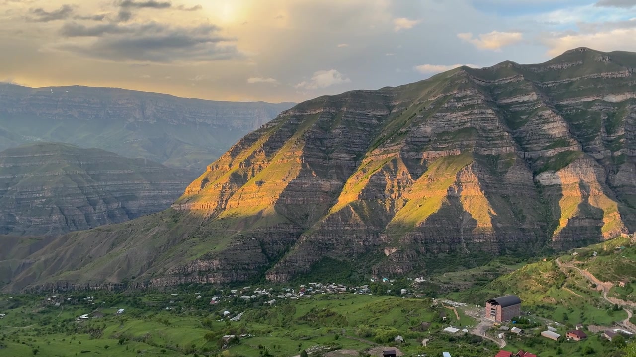La luce solare dell'ora d'oro illumina le montagne verdi stratificate del Dagestan, con un piccolo villaggio annidato nella valle sottostante sotto un cielo nuvoloso.