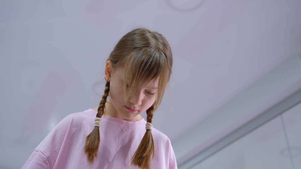маленький ребенок в розовом наряде с плетеными волосами, смотрящий вниз с сосредоточенным выражением лица в ярко освещенной комнате, возможно, занимающийся концентрацией или деятельностью