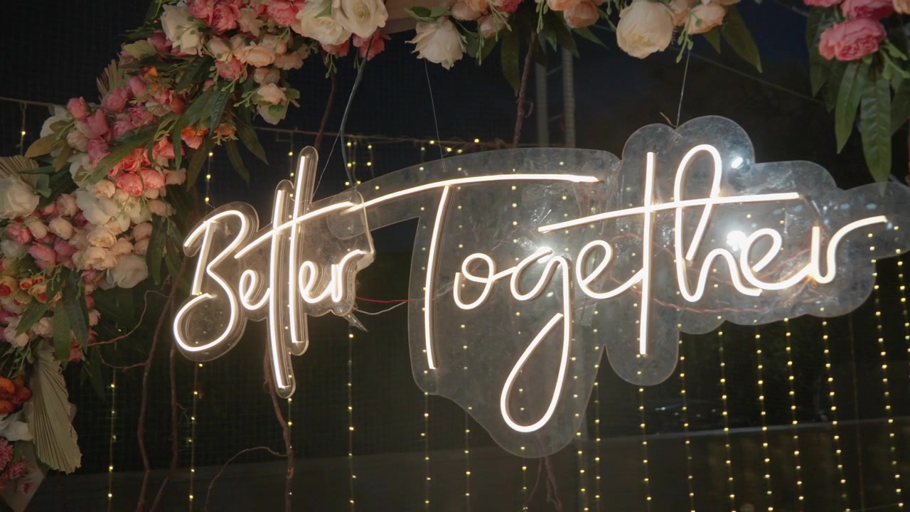 Insegna al neon luminosa "Better Together" incorniciata da fiori rigogliosi e lucine fatate a una celebrazione Sangeet dell'Asia meridionale.