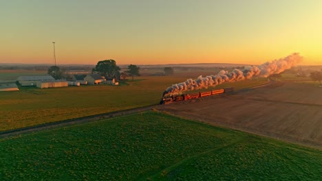 aerial view of a steam engine with passenger cars at the golden sunrise approaching with a full head of steam and smoke traveling thru the farmlands