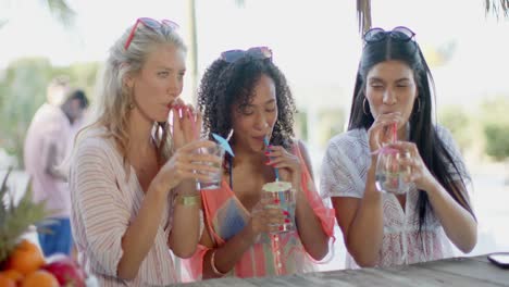 Happy-diverse-female-friends-drinking-cocktails-at-beach-bar-and-talking