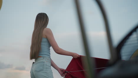 young woman in denim shorts holds red ripstop panel tether of colorful hot air balloon envelope while nearby engine blurs in foreground during breezy sunset field preflight setup scene