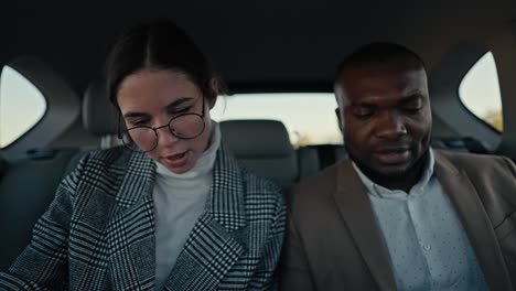 A-confident-brunette-girl.-A-businessman-with-round-glasses-in-a-gray-jacket-communicates-with-his-colleague-a-man-with-Black-skin-and-a-beard-in-a-brown-jacket-during-his-business-trip-in-the-cabin-of-a-modern-car-outside-the-city