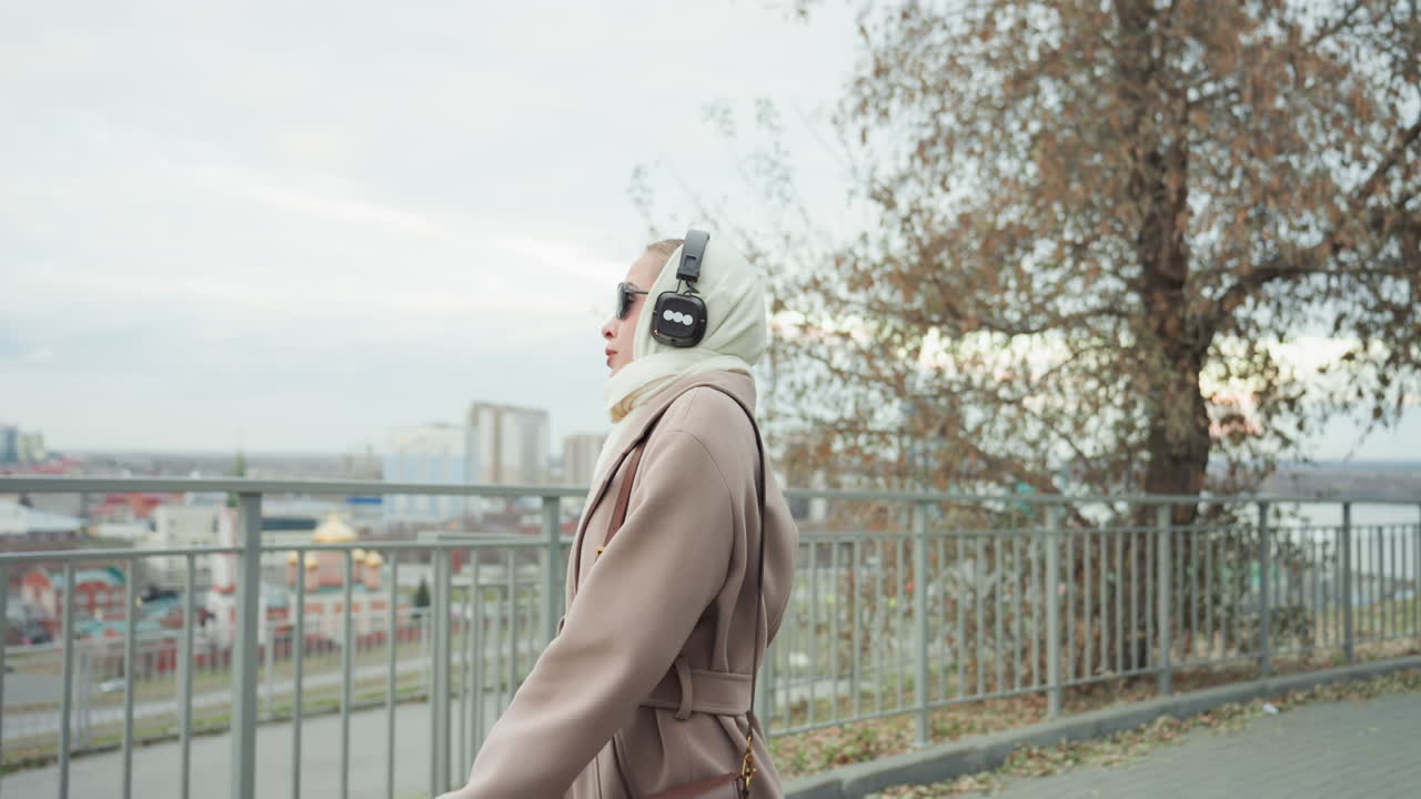 Kaukasisk kvinde går ved et rækværk med udsigt over byens skyline iført trenchcoat, håndtaske og høretelefoner, selvsikker gang, pendler, indholdsskaber, urban efterårsscene, let vind, udsigt over byen