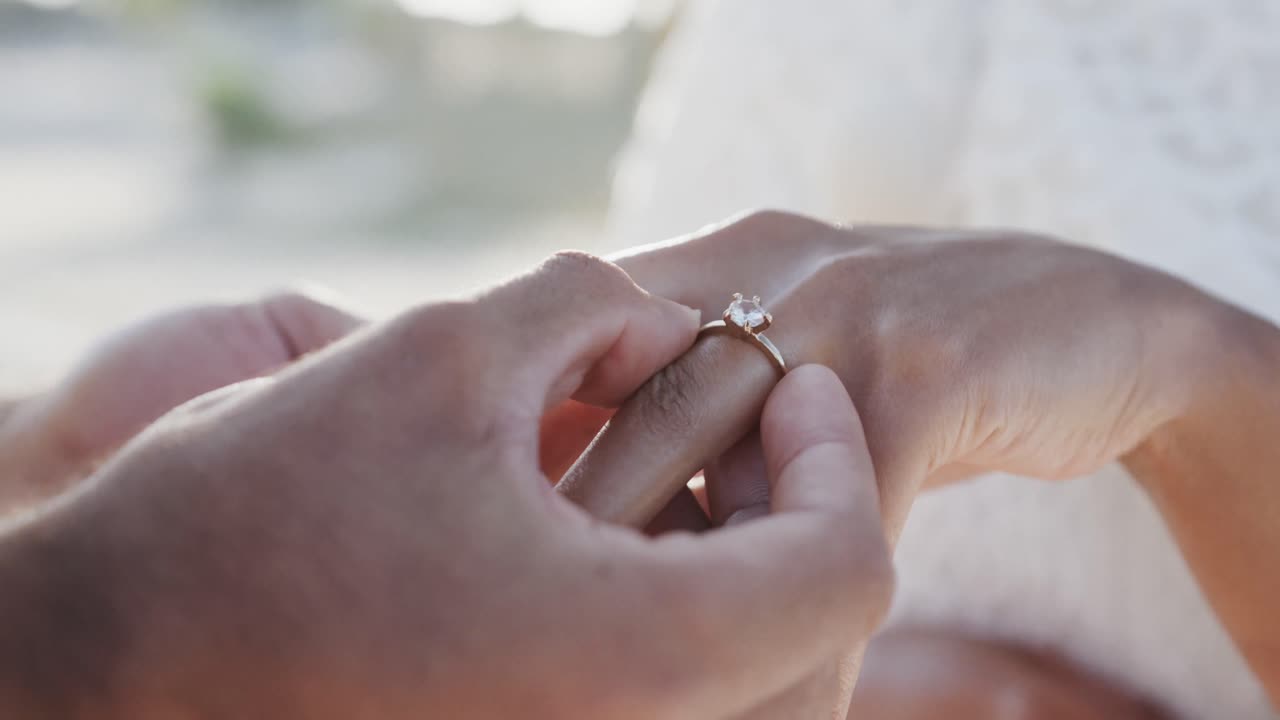 Hands of diverse groom putting wedding ring on finger of bride at beach wedding, in slow motion