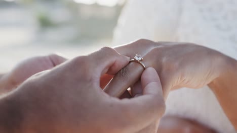 hands of diverse groom putting wedding ring on finger of bride at beach wedding, in slow motion