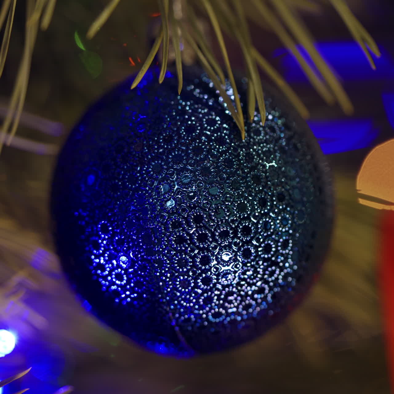 Traditional Christmas decoration on a spruce tree. Blue glass Christmas ball hanging on a pine tree on colorful spots background. New Year shiny bauble. Close-up.