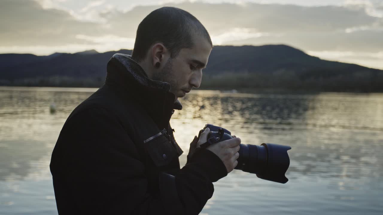 Young Caucasian man looking through and adjusts handheld camera standing by calm lake waters with dark mountain range in background, profile close up