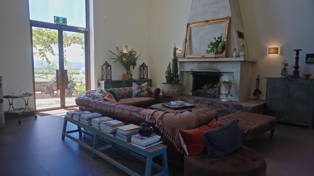 A cozy vintage living room with brown sofa, gold empty frame, books, green plants, and subtle wedding decor. Glass doors reveal a bright daytime beach view outside