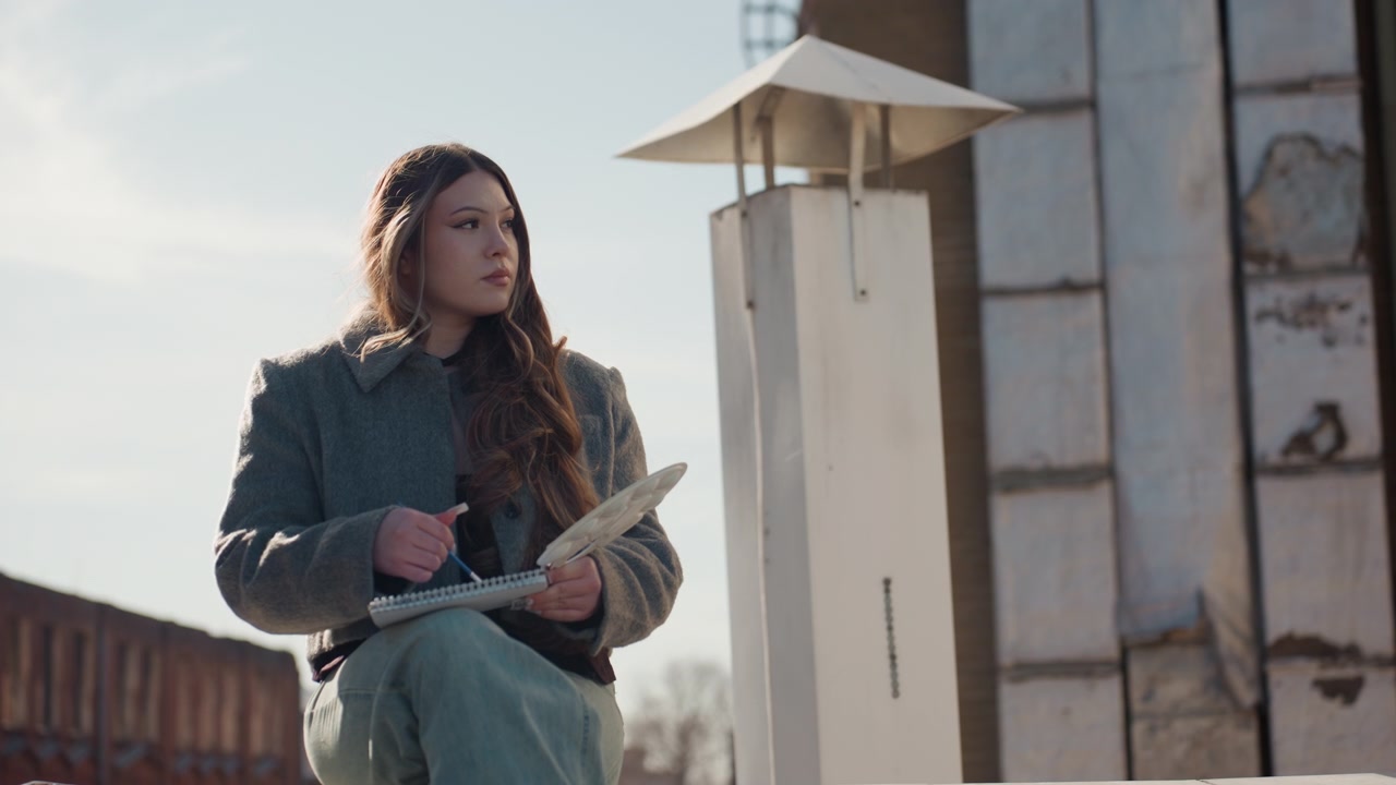Pretty woman on rooftop arranging dried wheat, textured coat, seated near industrial chimney, soft lateafternoon sunlight, gentle breeze, careful hands tying stems, muted cinematic palette