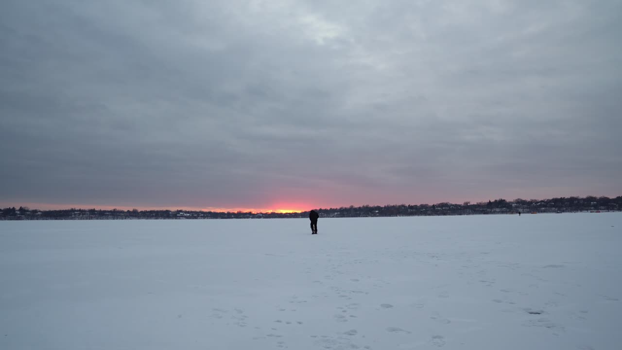 Man wearing headphones walks with his pug across a vast frozen lake in Minnesota during dusk