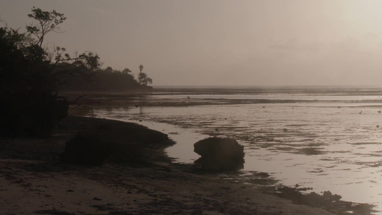 Local people walk across the exposed reef at low tide near G-Land, Indonesia, surrounded by warm sunset haze and soft reflections across the tropical coastline.