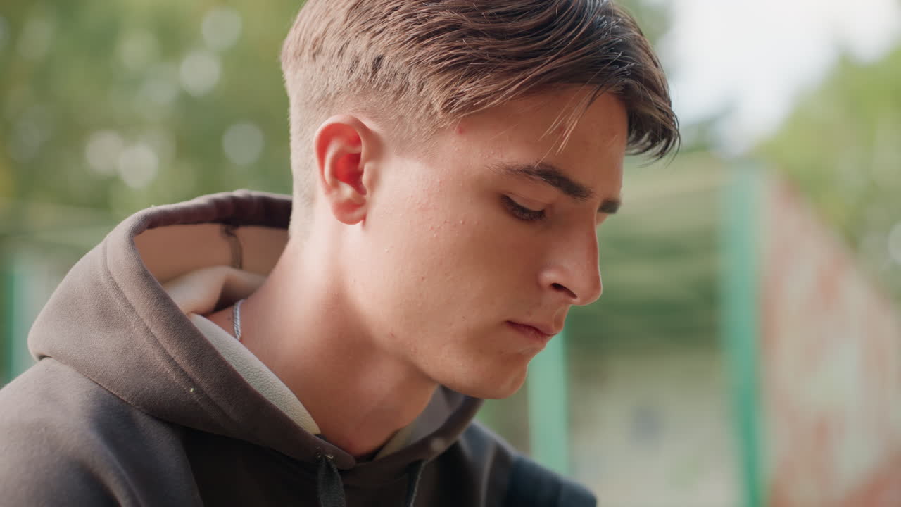 Adolescente con felpa con cappuccio che guarda in basso, ritratto cinematografico di un parco con espressione pensierosa, luce dorata soffusa e profondità di campo ridotta, studente che aspetta alla fermata dell'autobus, primo piano spontaneo che cattura la vulnerabilità