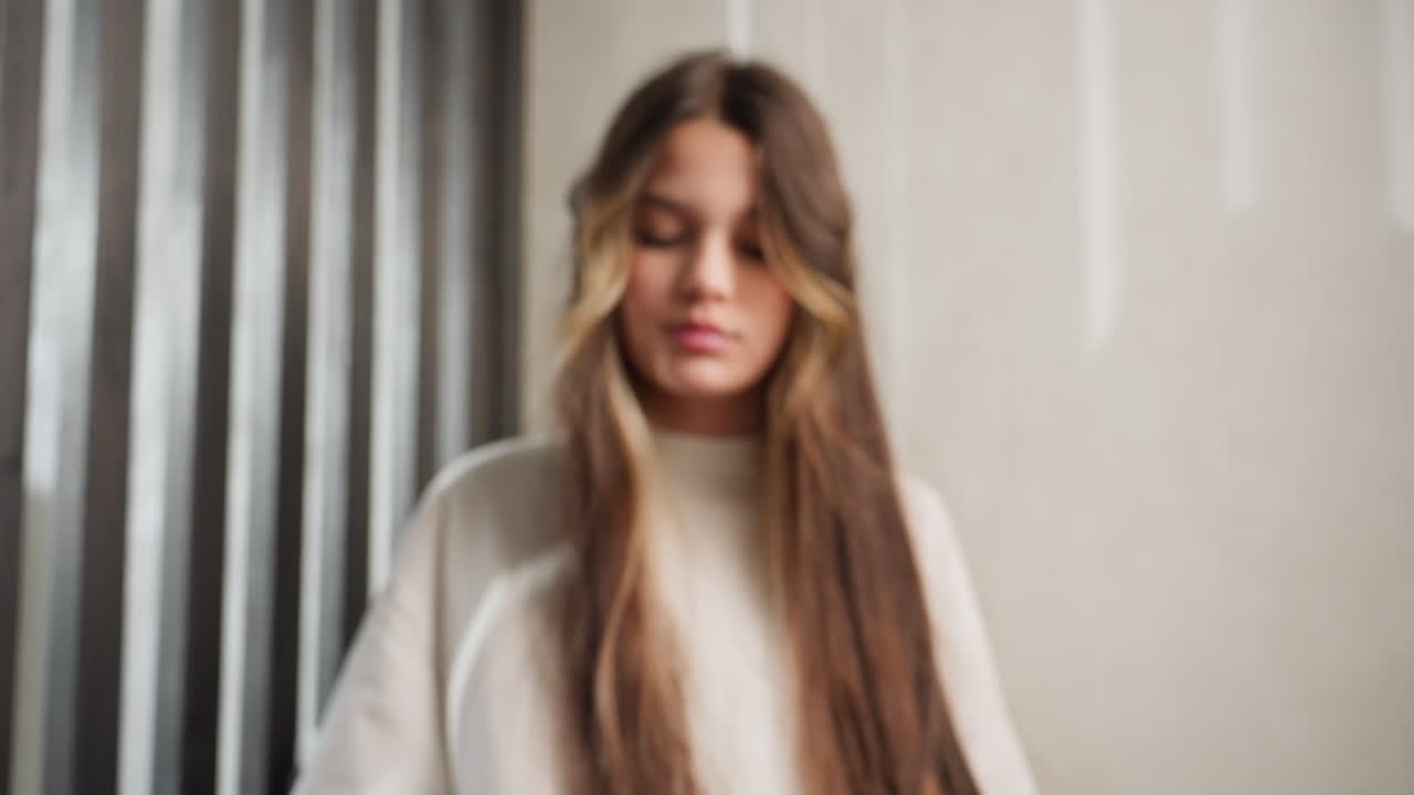 Woman with long hair enters studio corridor, walks to seat, gentle head turn, soft hair flip, sits gracefully, calm mood, ribbed wall panels, cinematic light, slow movement, subtle elegance