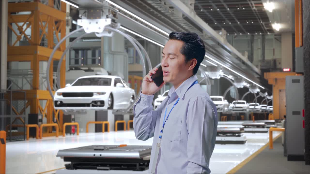 보기: 아시아 남성 직업 노동자가 ev 자동차 공장, 하이테크 산업 시설에 서서 전화를 들고 끔한 미소로 쳐다보기 시작했습니다.
