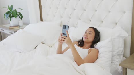 Glückliche-Asiatische-Frau,-Die-Im-Bett-Liegt,-Kopfhörer-Trägt-Und-Ihr-Smartphone-Benutzt,-In-Zeitlupe