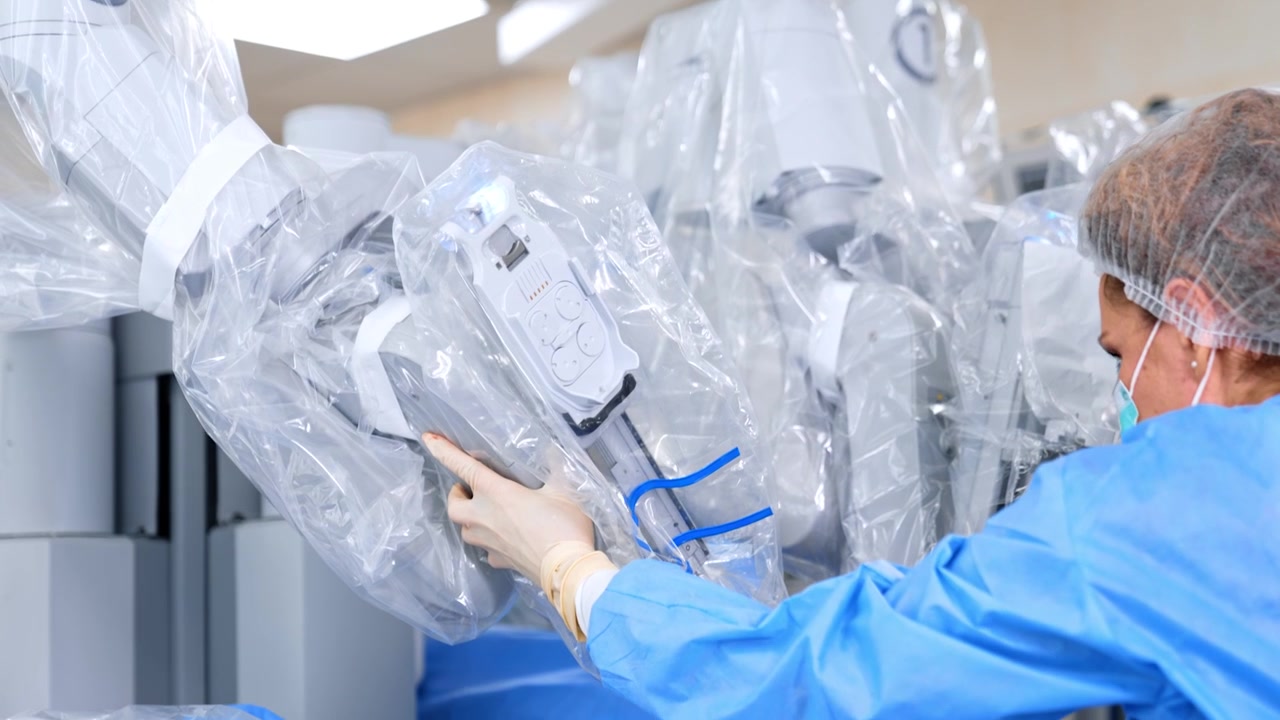Female doctor in mask, cap and blue robe pulls the robotic arm of surgery system closer to the patient. Da Vinci machine is covered in plastic for sterility.