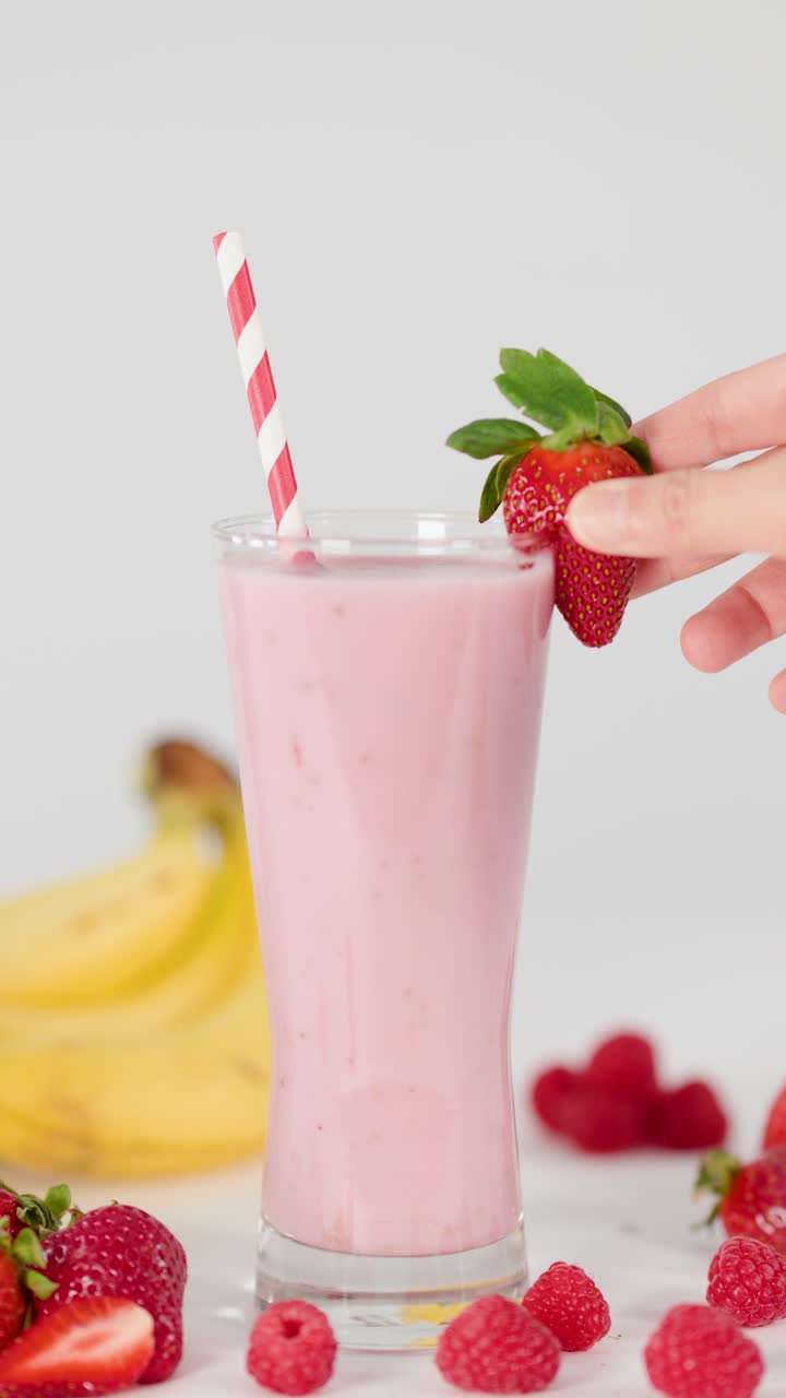 Mano añade un adorno de fresa a un batido rosa cremoso en vaso, rodeado de plátanos y frambuesas