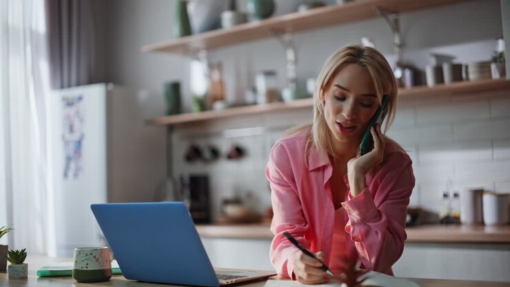 Donna multitasking che parla al cellulare mentre digita sul portatile in una cucina moderna, primo piano