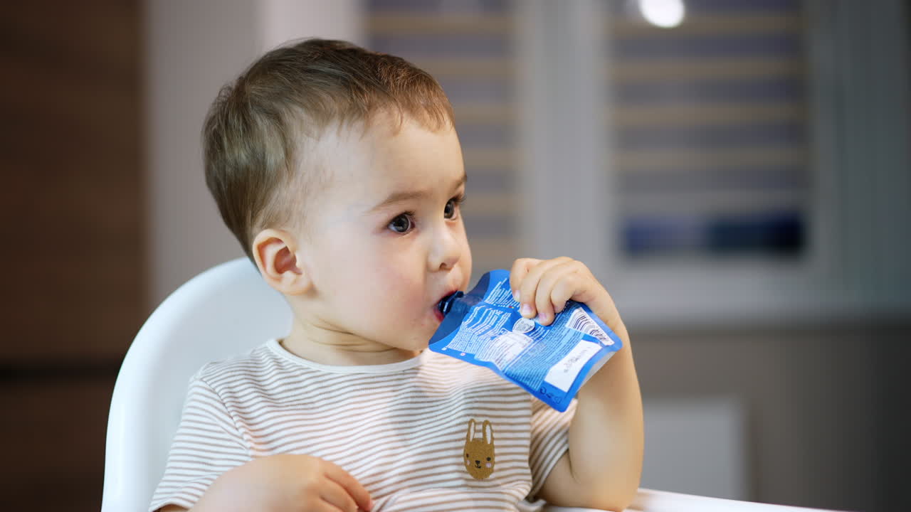 Caucasian toddler sitting in a high chair with a doy pack in hand. Adorable child fruit puree willingly. Close up.