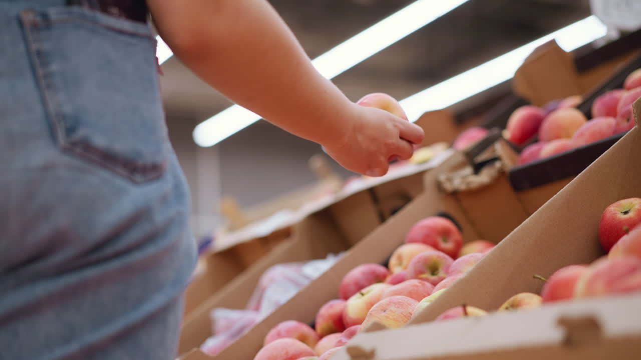 뒷 주머니에 드폰을 들고 사과에서 은 사과를 뽑고, 그것을 관찰하고, 활기찬 식료품 가게 설정으로 다시 떨어뜨리는 여자의 부분적인 시각