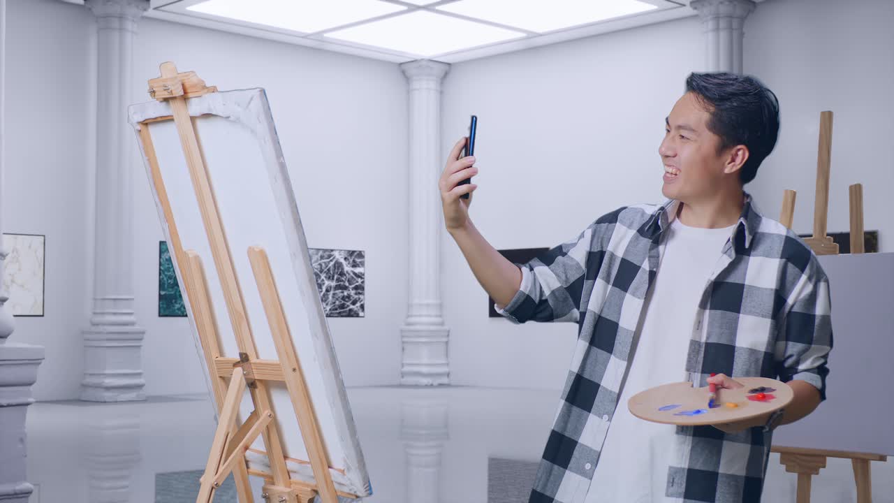 아시아 남성 예술가 가 갤러리 에서 오일 페인트 와 브러쉬 로 캔버스 에 그림 을 그리는 동안 스마트폰 으로 비디오 통화를 하고 있다