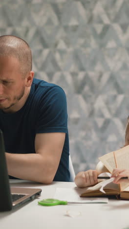 young daughter do homework, sits at table with book, on distance learning, dad sits next, works at computer and talks on phone, say girl not to interfere and not to distract