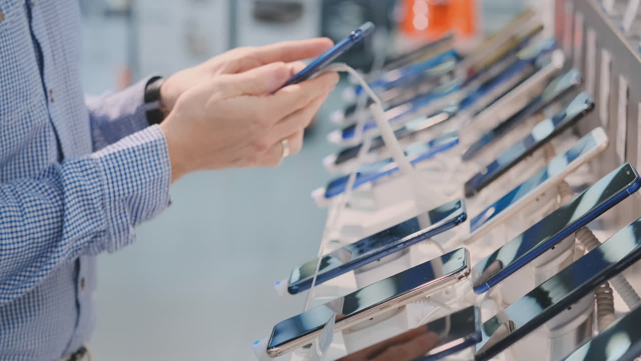 Close-up of a man's hand rotates and inspects the design of a new smartphone in a modern electronics store. Take the phone before buying
