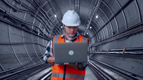 asian male engineer with safety helmet working on a laptop while standing in underground subway tunnel