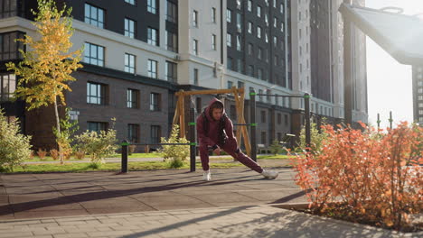 long shot of graceful woman in hoodie, vest performing side lunge workout with hands on legs on tile ground in urban courtyard framed by autumn foliage, bars and high tower under warm sunlight