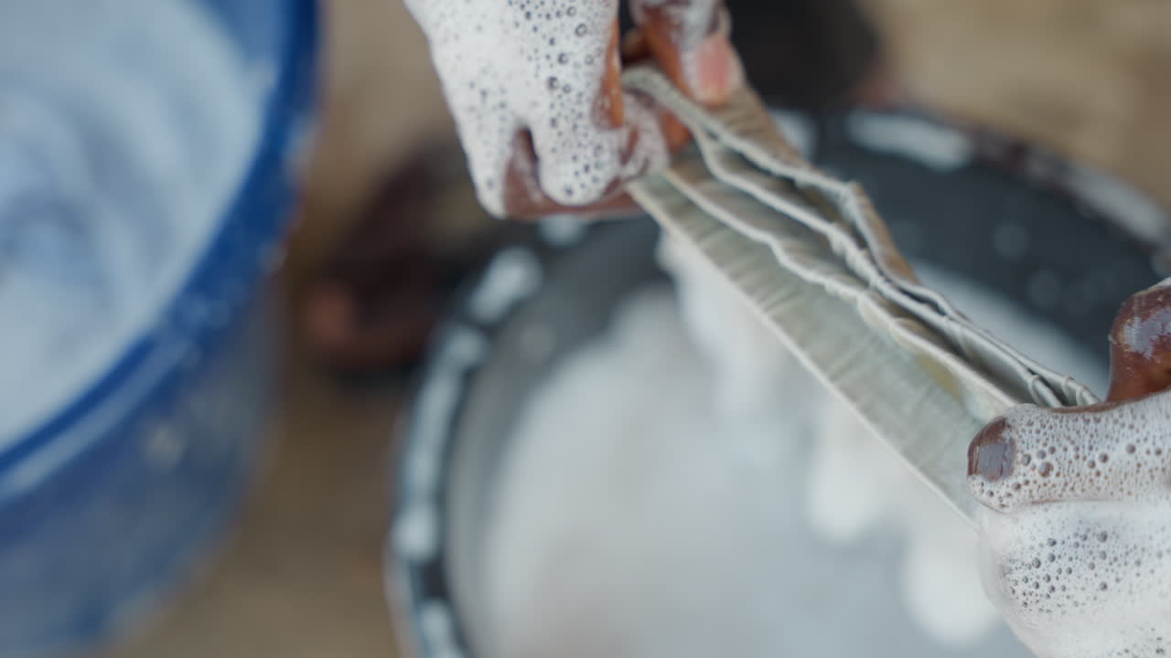 Black hands washing sock in bucket, heavy soap suds, blue plastic tub, outdoor concrete floor, homemaker scrubbing with steady rhythm, household chores captured in tactile closeups, water dripping