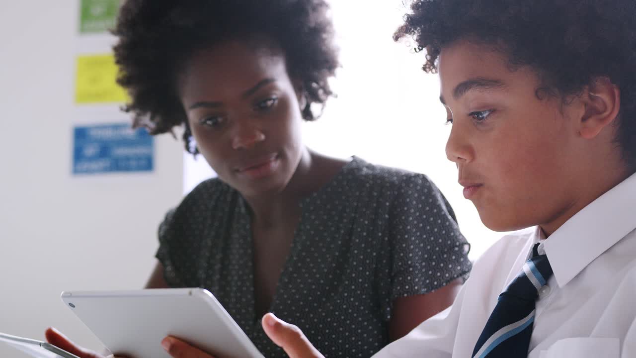 Close Up Of Female High School Tutor With Male Student Wearing Uniform Using Digital Tablets For One To One Tuition In Classroom
