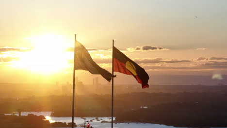 Bandera-Australiana-Y-Aborigen-Ondeando-Al-Atardecer,-Sydney-Al-Fondo,-Hora-Dorada,-Cinematográfica,-Aérea-Gran-Angular,-Toma-Circulante